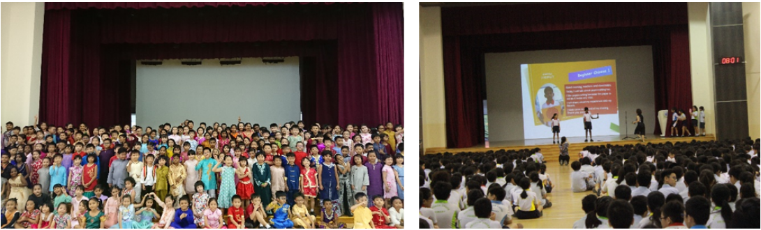 Large group of children in colorful traditional outfits pose on a stage, beside a school assembly with students watching a presentation.